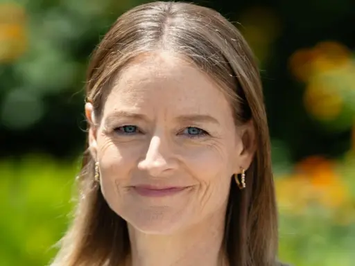 Frau mit blauen Augen und braunem Haar, die lächelt. Ein dezenter Ansatz von grauen Haaren wird durch die Frisur kaschiert.