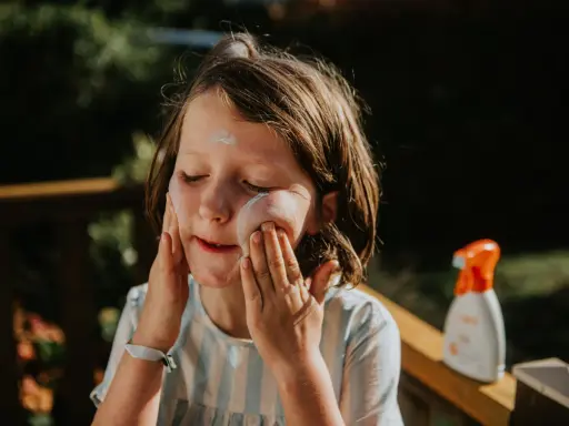 Kind mit geschlossenen Augen trägt Sonnencreme auf. Eine Flasche abgelaufene Sonnencreme steht daneben.
