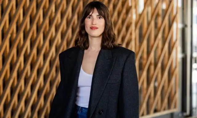 Frau mit französischem Crop-Haarschnitt, trägt einen Oversized-Blazer und Jeans vor einer Holzwand.