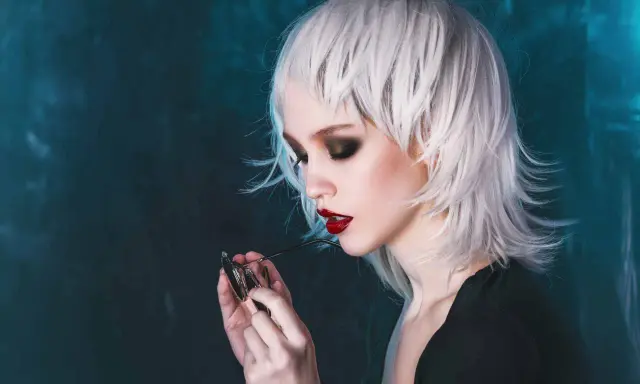 Stylische Dame mit weißem Wolfcut, dunklem Lidschatten und roten Lippen hält eine Brille.