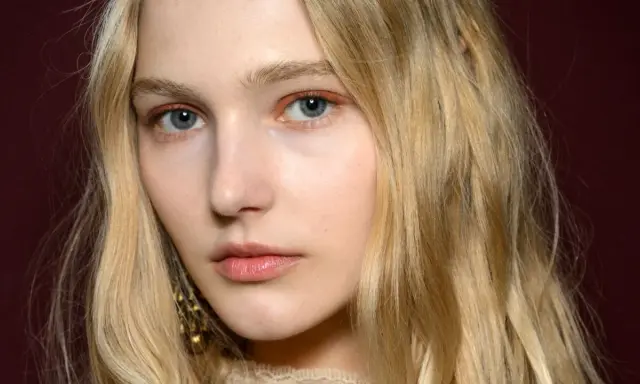 Blonde Haare umrahmen ein Gesicht mit blauen Augen und dezentem Make-up. Ihre hooded eyes wirken geheimnisvoll.