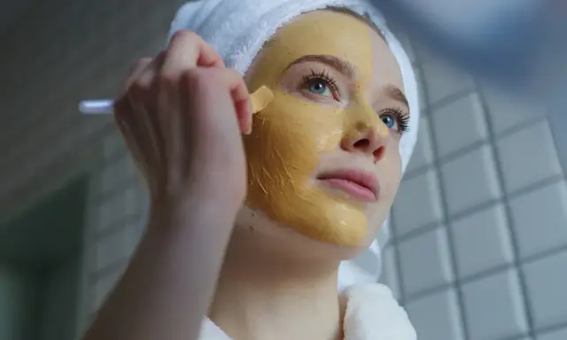 Junge Frau mit Handtuch auf dem Kopf trägt eine Gesichtsmaske auf. Die Kurkuma Wirkung auf die Haut ist bekannt für ihre entzündungshemmenden Eigenschaften.