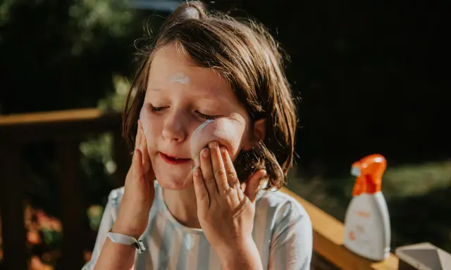 Kind mit geschlossenen Augen trägt Sonnencreme auf. Eine Flasche abgelaufene Sonnencreme steht daneben.