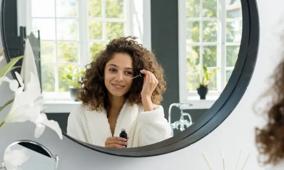 Frau mit lockigem Haar trägt ein Serum auf, um die Vitamin C Haut Wirkung zu erzielen. Sie lächelt im Spiegel.