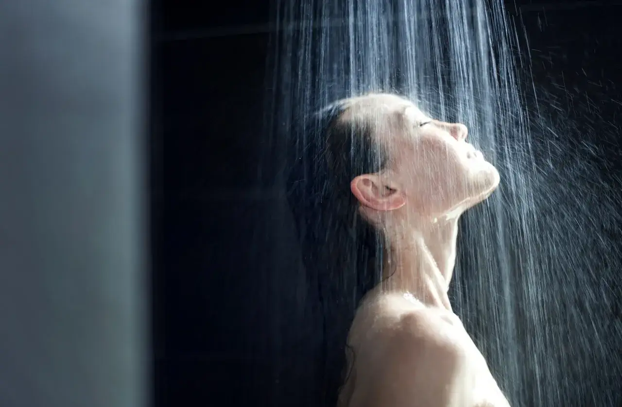 Frau genießt eine erfrischende Dusche. Das Wasser fällt auf ihr Gesicht, während sie die Augen schließt und sich dem Moment hingibt. Ein Gefühl von Reinheit und Entspannung beim richtig duschen.