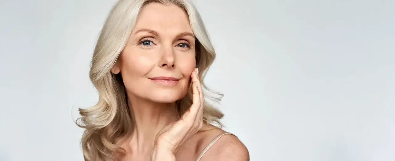 Frau mit blonden Haaren, die sanft ihr Gesicht berührt. Tipps, wie man Concealer richtig auftragen bei Falten kann, um jugendliche Haut zu zaubern.