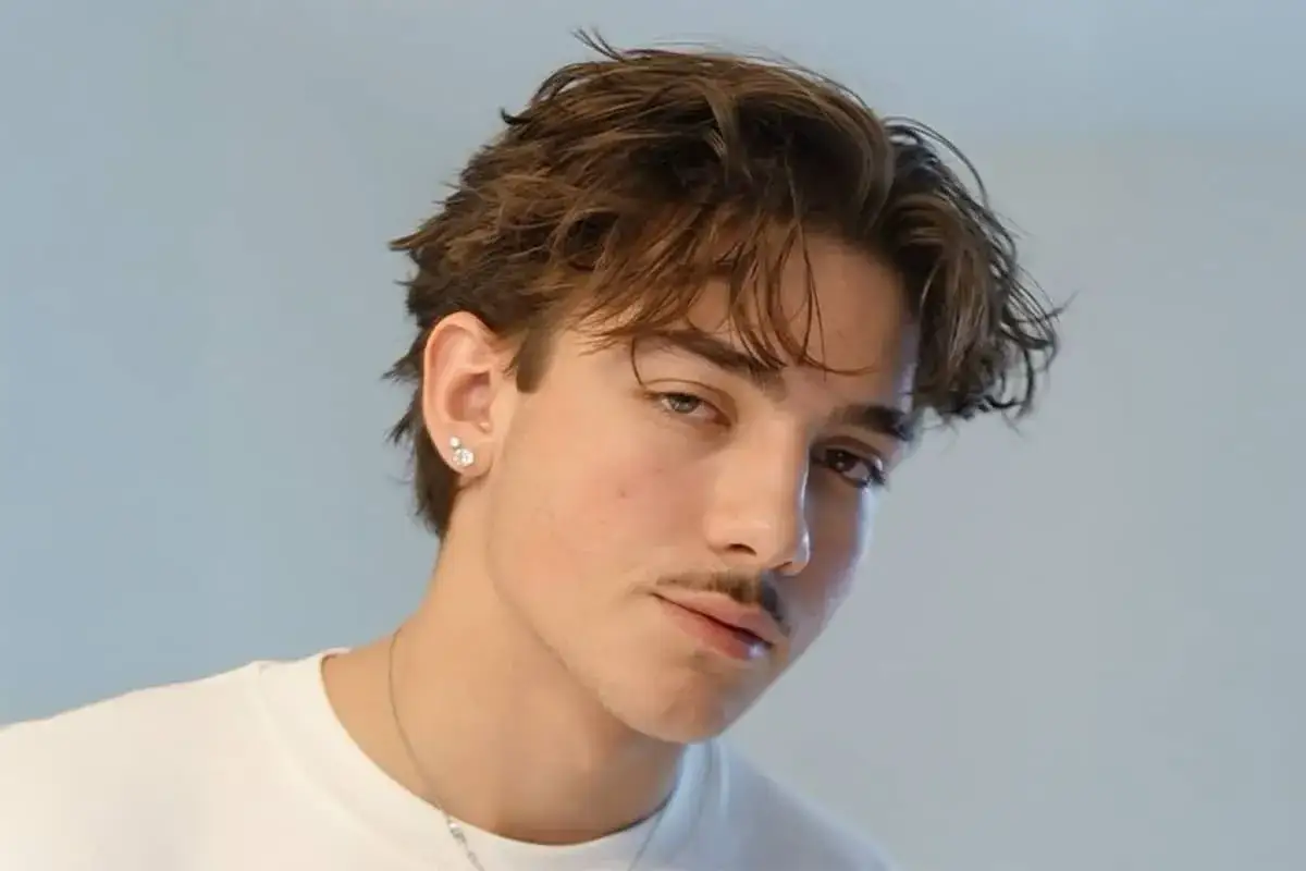 Junger Mann mit braunen Haaren und einem dezenten Schnurrbart, der einen Mittelscheitel trägt. Er hat einen Ohrstecker und trägt ein weißes T-Shirt.